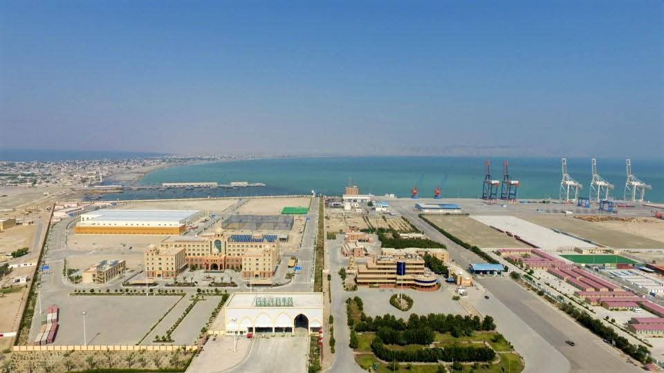 An aerial view of Gwadar Port with its cranes and facilities, alongside modern buildings and a structured road network, with the sea in the background.