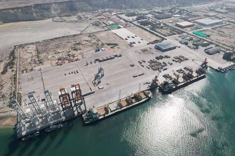 Arial view of Gwadar Port in Balichistan, Pakistan, featuring cargo cranes, docked ships, and port infrastructure along the waterfront, with urban development visible in the background